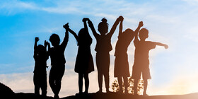 A group of kids holding hand and raising their arms, backlit against a sunset.