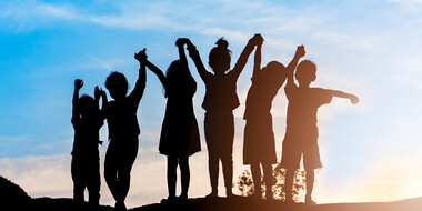 A group of kids holding hand and raising their arms, backlit against a sunset.